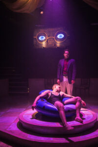 Production Photo from The Great Gatsby: Jay Gatsby lies limp on an inner tube; Nick Carraway stands over him. The eyes on the billboard glow behind him.